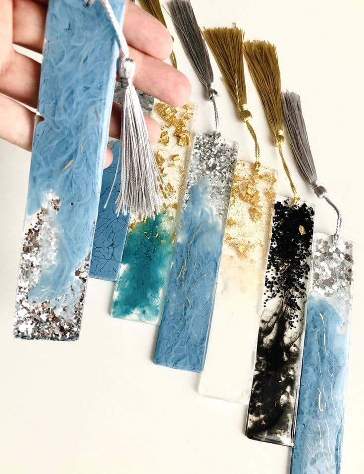 En hånd holder et fantastisk harpiksbokmerke med en sølvkvast, laget med Glassklar Epoxy - CCR Epoxy Entropy SuperSap. Fem andre harpiksbokmerker vises på en hvit overflate, hver utsmykket med gylne eller grå dusker. Bokmerkene viser frem ulike design med marmorering og glitter i nyanser av blått, hvitt, gull og svart.